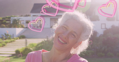 Smiling senior woman wearing pink top standing on concrete steps of white house, with heart icons....