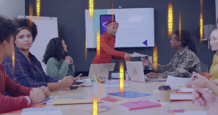 Presenter in red turtleneck and headband handing document in meeting room with laptops and screen....