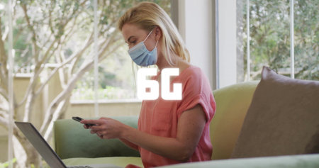 Woman wearing coral pink shirt, surgical mask, using smartphone and laptop on sofa by garden window. Lifestyle, relaxation, modern, home, comfort, technology, serenityの写真素材