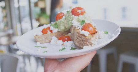 Hand holding plate in cafÃ©, with avocado toast, halved tomatoes, crumbled white cheese, cilantro....