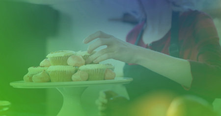 Placing woman in red top and apron arranging frosted cupcakes on cake stand on kitchen table. Bakery, baking, confections, desserts, culinary, artisan, home decorの写真素材