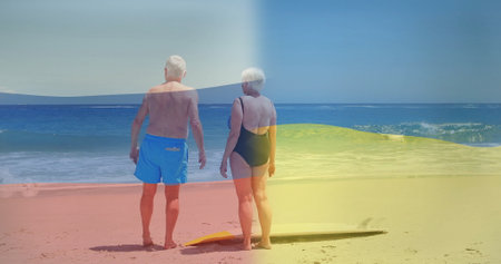 Standing senior couple gazing at ocean on sandy beach, with yellow bodyboard nearby. Sunset,...