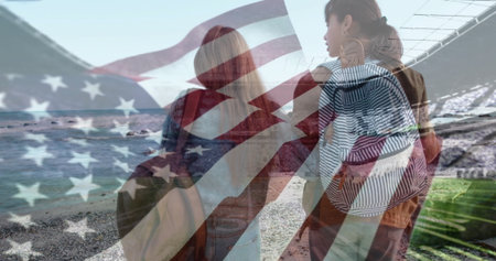 Standing teen girls facing ocean waves lapping shore, with flag, striped backpack and tote bag. Adventure, friendship, youthful, freedom, exploration, coastal, patrioticの写真素材