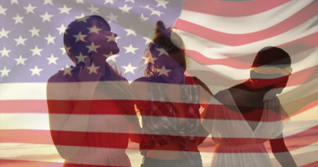 Gazing three friends linking arms on sandy beach at sunset, with translucent US flag overlay. Friendship, unity, celebration, harmony, leisure, serenity, diversityの写真素材