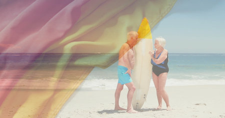 Standing senior couple wearing swim trunks and one-piece facing ocean, with yellow-tipped surfboard. Adventure, relaxation, leisure, coastal, outdoor, carefree, vitalityの写真素材