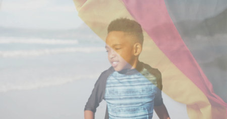 Waving multicolored towel overhead boy wearing blue swim shirt standing on sandy beach, ocean waves....