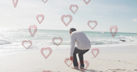 Bending senior Hispanic man picking up pink flamingo stake on beach, with floating heart overlay. Relaxation, coastal, serenity, wellness, mindfulness, casual, vibrantの写真素材