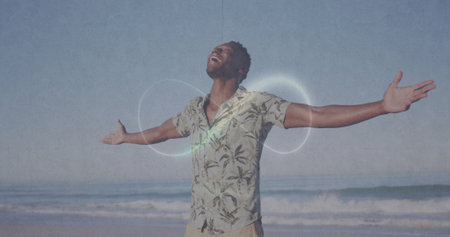 Spreading arms man wearing short-sleeve palm-print shirt smiling at sandy shore with ocean waves....