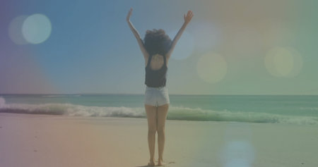 Raising arms, African American woman facing ocean on shore, wearing black tank top and blue shorts....