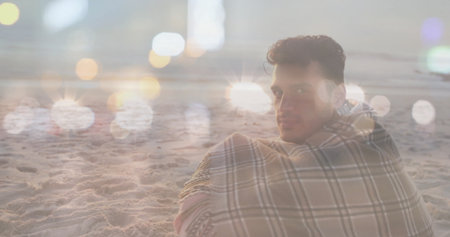 Sitting Hispanic man wrapped in plaid blanket on beach at night, with headlight bokeh, copy space. Solitude, relaxation, nighttime, serenity, reflective, cozy, outdoorの写真素材