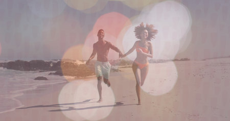 Running couple wearing orange bikini, trunks on sandy shore, with waves crashing on rocks. Vacation, leisure, freedom, vibrant, outdoors, adventure, summerの写真素材