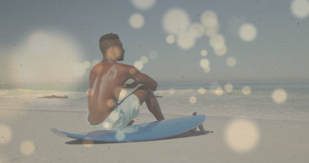 Sitting shirtless man wearing swim trunks gazing toward ocean on beach, with blue surfboard. Seaside, adventure, outdoor, leisure, tranquility, fitness, sunlitの写真素材