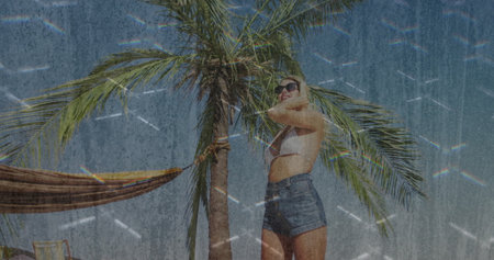 Standing woman wearing sunglasses, bikini top, shorts on beach by palm, striped hammock, copy space. Tropical, leisure, relaxation, lifestyle, minimalism, outdoor, sunnyの写真素材