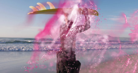 Walking surfer wearing black wetsuit holding surfboard overhead on beach, with swirling pink powder....