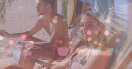 Relaxing couple wearing trunks and bikini top sitting on beach chairs under palms, with surfboard....