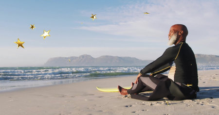 Sitting surfer in wetsuit gazing at sea on beach with yellow surfboard, star graphics, copy space. Adventure, leisure, sport, coastal, vibrant, inspirational, outdoorの写真素材