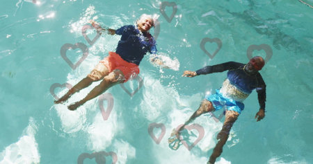 Floating senior couple wearing rash guards and trunks in backyard pool, with heart overlays. Relaxation, tranquility, wellness, leisure, harmony, reflection, aquaticの写真素材