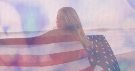 Standing woman holding draped American flag at beach shoreline, with ocean waves and pastel sky. Patriotism, freedom, serenity, nature, inspiration, landscape, celebrationの写真素材