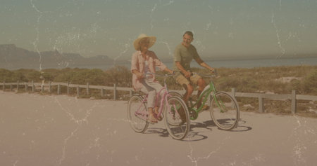 Cycling couple riding pastel cruiser bicycles on seaside path past dunes with straw hat and fence....
