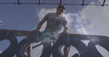 Climbing man wearing grey t-shirt black shorts gripping beam over strapped tires on obstacle course....