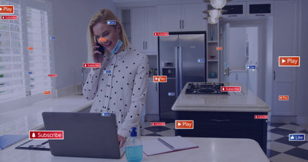 Talking mid adult woman in polka dot blouse leaning over laptop at kitchen island, with smartphone....