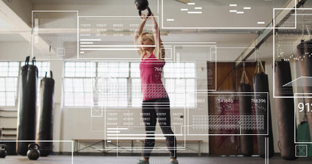 Mid-adult woman in pink top lifting heavy kettlebell overhead in gym, punching bags, data overlays. Fitness, strength, training, workout, athleticism, digital, modernの写真素材