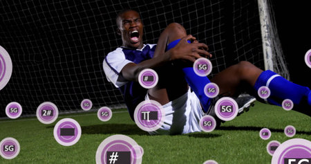Clutching right knee soccer player yelling on turf at night, with goal net, floating 5G icons. Athletic, motion, outdoor, sport, technology, digital, determinationの写真素材