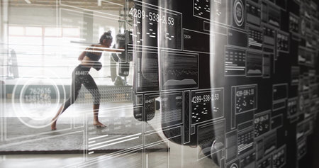 Female trainee in sportswear punching heavy bag on workout mat in gym, with digital data panels....