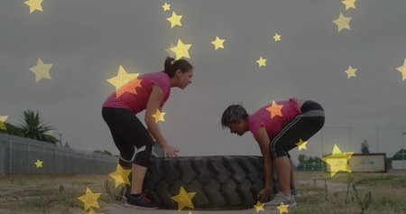 Friends in pink tops and black leggings flipping large tractor tire on flat dirt, chain-link fence....