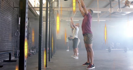 Working four people in gym clothes throwing overhead medicine balls in fitness gym, with rigs....