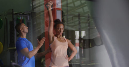 Female client performing kettlebell overhead press in gym, with trainer and weight rack. Fitness,...