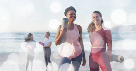 Smiling four women in athletic wear holding rolled yoga mats, chatting on sandy beach water's edge. Flexibility, outdoor fitness, wellness, camaraderie, summer activity, health, leisureの写真素材