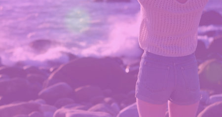 Standing woman wearing knit top, jean shorts on pebble shore, with breaking waves, copy space. Coastal, leisure, minimalist, tranquility, natural, outdoor, fashionの写真素材