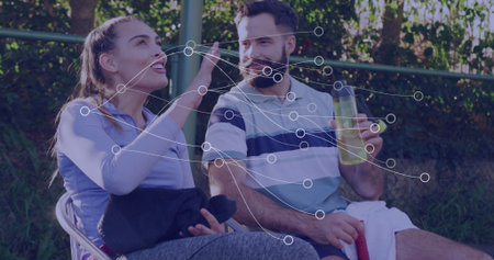Sitting tennis couple in sportswear chatting on chairs beside fence and hedge, holding water bottle. Athletic, leisure, casual, outdoor, vibrant, teamwork, relaxationの写真素材