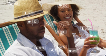 Relaxing couple wearing straw hats holding drink on deck chairs on sand, with floating social icons. Vacation, leisure, serenity, lifestyle, seaside, outdoor, tranquilityの写真素材