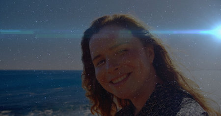 Smiling woman wearing patterned dark shirt gazing at ocean shoreline, under bright lens flare....