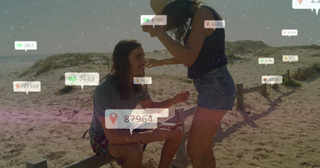Showing smartphone by woman toward man on sandy beach wooden fence, with notification overlay icons. Coast, relaxation, communication, leisure, outdoors, tranquility, lifestyleの写真素材