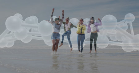 Leaping four women raising arms on wet sand with water lapping ankles, with molecular overlay. Joy, camaraderie, vitality, unity, movement, nature, healthの写真素材