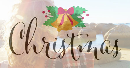 Standing woman in red-black coat holding smartphone on sandy beach, with Christmas bells graphic. Coastal, winter, festive, casual, leisure, scenic, vibrantの写真素材