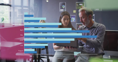 Reviewing laptop, two colleagues wearing business attire working at office with bar chart overlay....