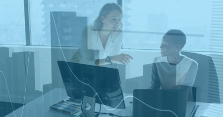 Collaborating women in business attire reviewing documents in office, with laptop, monitors....
