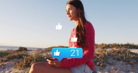 Sitting woman in sweater and shorts holding smartphone on dune viewing thumbs up icons, copy space....