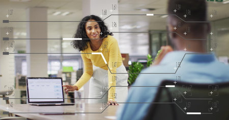 Presenting woman in yellow top leaning over table in open-plan office, with laptop data chart. Collaboration, teamwork, modernoffice, businessmeeting, corporateenvironment, productivity, professionalstyleの写真素材
