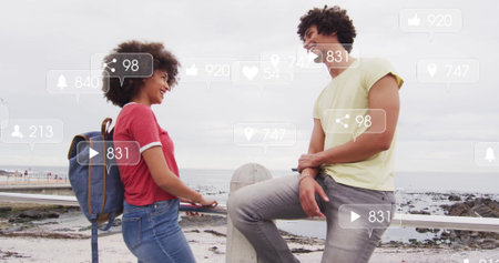 Leaning couple on metal railing by rocky shoreline, with blue backpack displaying social icons. Coastal, engagement, adventure, leisure, modern, waterfront, connectivityの写真素材