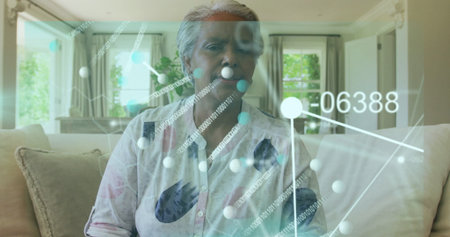 Sitting senior woman wearing shirt showing network overlay on home sofa with curtains potted plant....