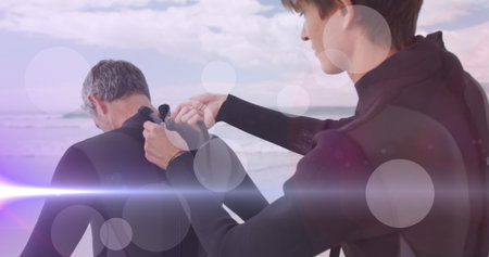 Younger man pulling zipper on older friend's wetsuit at beach shoreline, with full-body wetsuits. Adventure, camaraderie, outdoor, ocean, sport, teamwork, coastalの写真素材