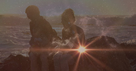 Sitting couple watching gentle waves at large coastal rock, with lens flare and starry sky. Nature,...