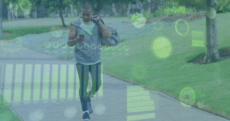 Walking athletic man glancing at smartphone on park path, with duffle bag, headphones and HUD....