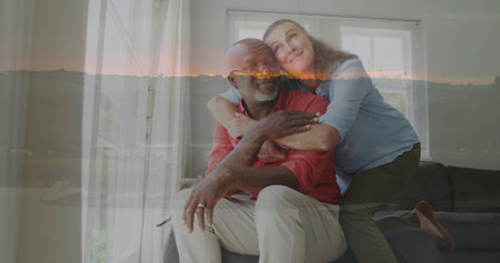 Hugging senior man and woman in living room on grey sofa by window with white curtains....