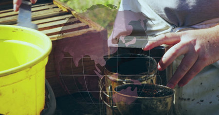 Operating beekeeper in grey zip-up top using bee smoker and hive tool at apiary with bucket. Apiary, beekeeping, agricultural, nature, rustic, outdoor, craftsmanshipの写真素材